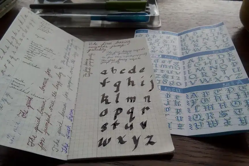 An open notebook of handwriting practice beside a printed alphabet card on a wooden desk