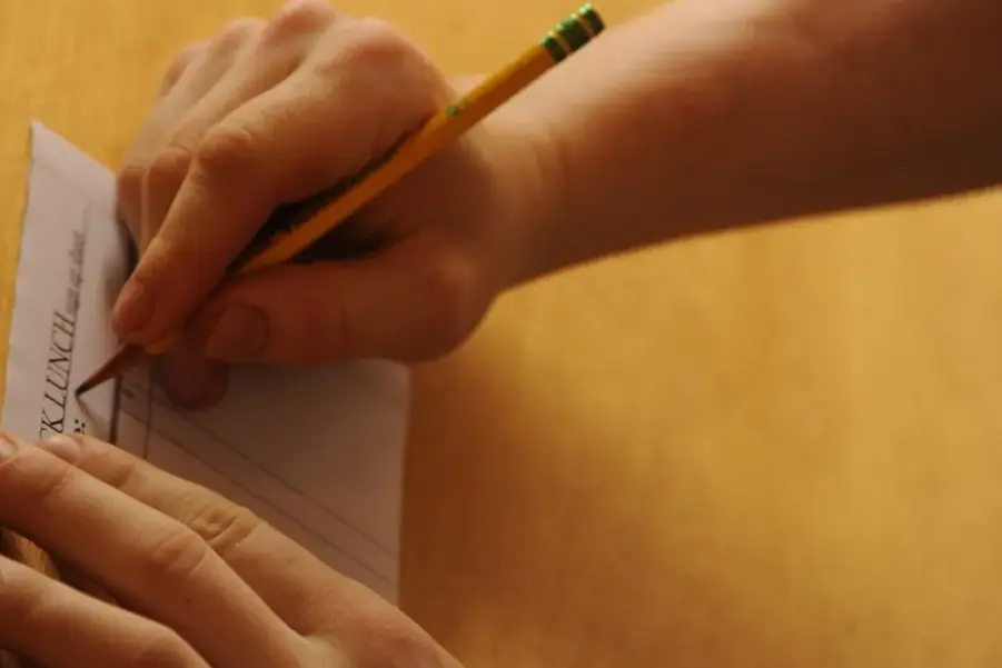 Two hands holding a sheet of ruled paper steady while a yellow pencil writes on it